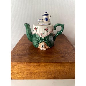 May include: A ceramic teapot with a green handle and spout, and a white base with a green curtain design. A small teapot and cup sit on top. The teapot features a sleeping dog face at the base.
