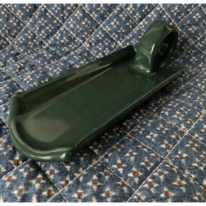 May include: A dark green ceramic soap dish with a curved design and a handle. The dish has a glossy finish and is set against a blue and white patterned background. The dish is approximately 8 inches long.