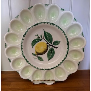 May include: A white ceramic oyster serving platter with a central bowl featuring a lemon branch design. The outer ring has 12 oval compartments with a pale green hue, and a green scalloped edge. The platter is displayed on a gold-colored metal stand.