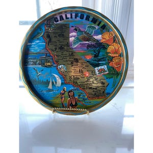 May include: A round decorative tray featuring a map of California with gold accents. The tray has a blue background with images of a quail, poppies, and a sailboat. The word "CALIFORNIA" is at the top.