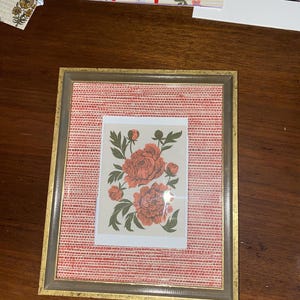 May include: A framed floral print featuring coral-colored peonies with green leaves. The print is set against a red woven background within a gold-toned frame. The frame is rectangular and has a neutral color.