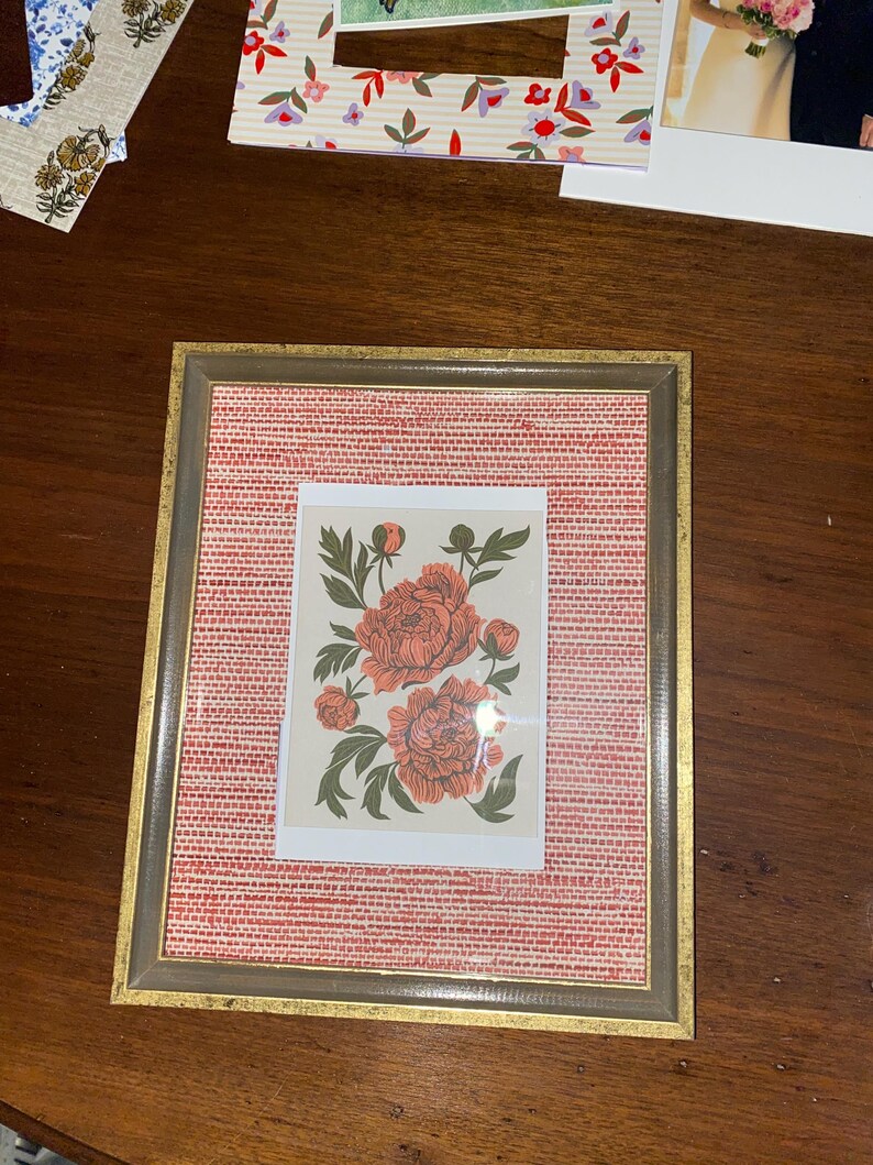 May include: A framed floral print featuring coral-colored peonies with green leaves. The print is set against a red woven background within a gold-toned frame. The frame is rectangular and has a neutral color.