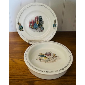 May include: Two white ceramic plates with illustrations of Peter Rabbit. One plate is round, the other is a shallow bowl. Text encircles the rims. The illustrations depict scenes from the Peter Rabbit story, with colorful details.