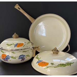 May include: Cream-colored enamel cookware set featuring a skillet and two lidded pots. The set is adorned with floral designs in orange, blue, and purple, with gold-toned handles and rims. A vintage kitchenware set.