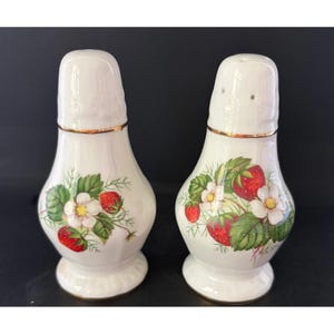 May include: A pair of white ceramic salt and pepper shakers featuring a botanical strawberry pattern with red berries, green leaves, and white blossoms. Each shaker has a gold-toned rim near the top and a flared base with a smooth, glossy finish.