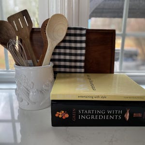 May include: A collection of kitchen utensils, including wooden spoons, a spatula, and a whisk, are displayed in a white ceramic utensil holder. Two cookbooks, one with a yellow cover and the other with a black cover, are stacked in front of the utensils.