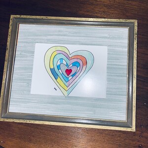 May include: A framed artwork featuring a colorful heart design. The heart is composed of concentric layers in various colors, including red, blue, yellow, and orange. The artwork is signed "Mia" and is set within a gold-toned frame.