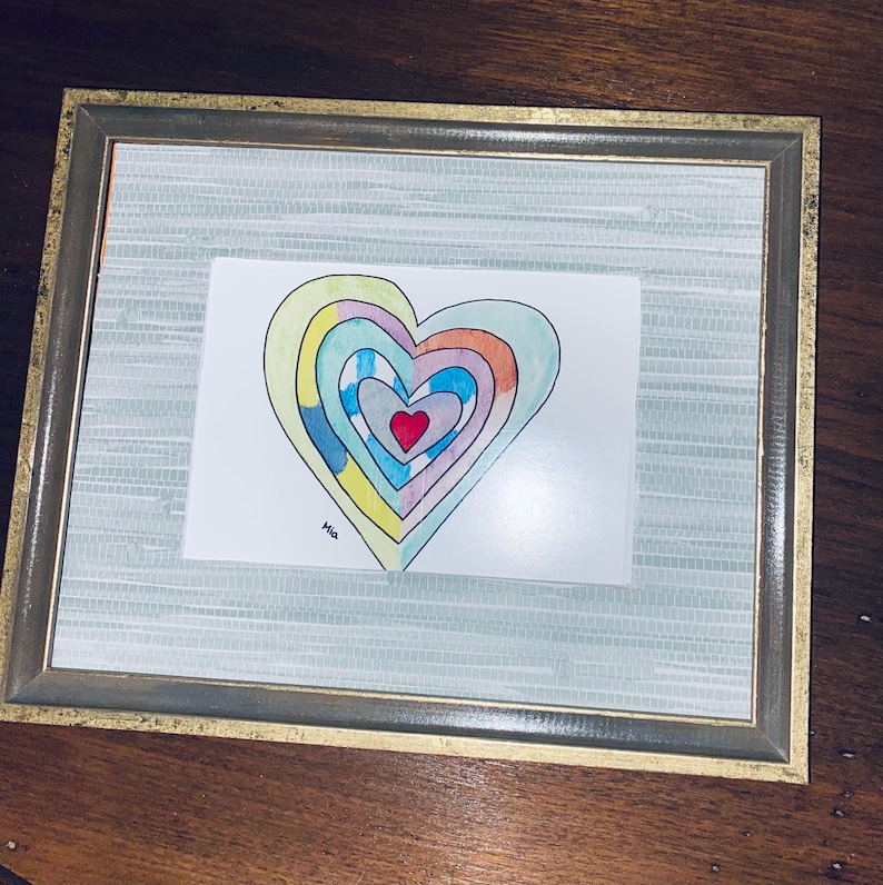 May include: A framed artwork featuring a colorful heart design. The heart is composed of concentric layers in various colors, including red, blue, yellow, and orange. The artwork is signed "Mia" and is set within a gold-toned frame.