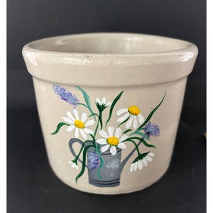 May include: A beige ceramic planter featuring a hand-painted design of a grey watering can filled with white daisies with yellow centers and purple lavender sprigs. The pot has a smooth, glossy finish and a wide, rounded rim at the top edge.