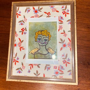 May include: Framed artwork featuring a whimsical angel illustration with yellow hair and golden wings. The artwork is set against a floral patterned background with a gold-toned frame. The angel has a green body and rosy cheeks.