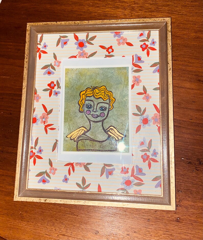 May include: Framed artwork featuring a whimsical angel illustration with yellow hair and golden wings. The artwork is set against a floral patterned background with a gold-toned frame. The angel has a green body and rosy cheeks.