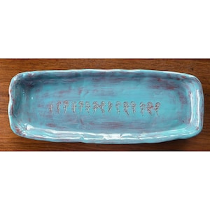 May include: A rectangular ceramic tray with an uneven, wavy rim. The surface is glazed in a vibrant turquoise blue with visible reddish-brown brushwork and a central horizontal row of small, stamped floral motifs in a dark brown tone.