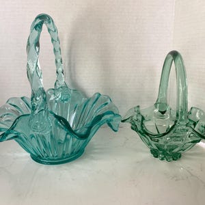 May include: Two aqua-colored glass baskets. The larger basket has a braided handle and a ruffled rim. The smaller basket has a curved handle and a scalloped rim. Both baskets are made of clear glass.