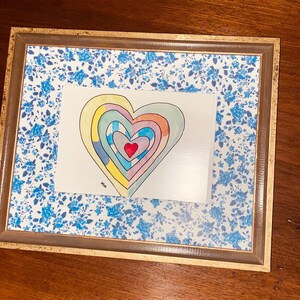 May include: A framed artwork featuring a colorful heart design. The heart is composed of concentric layers in various colors, including red, yellow, and blue. The frame has a gold-colored trim and a blue floral patterned background.