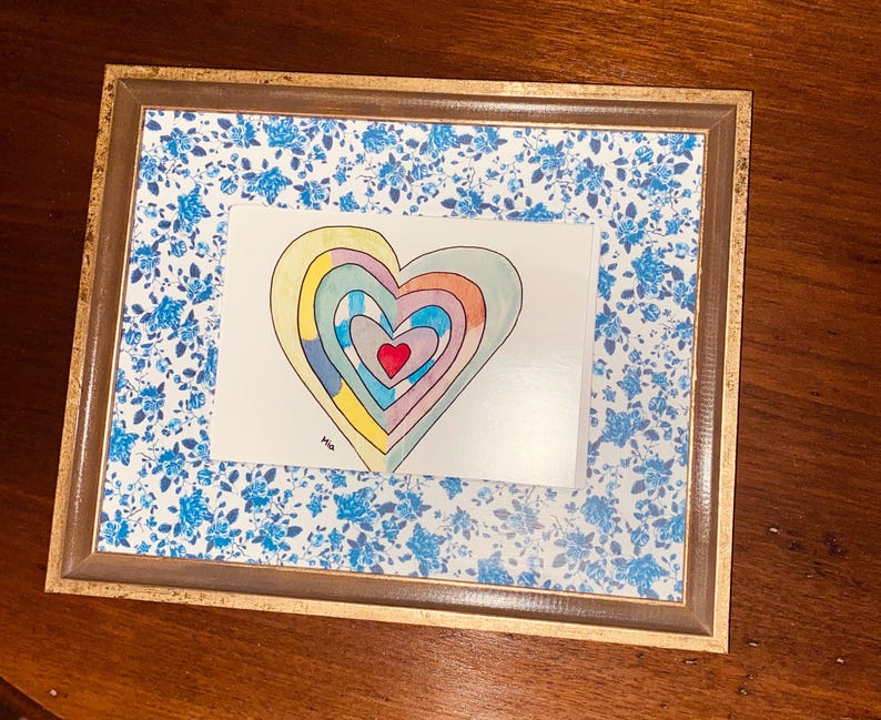 May include: A framed artwork featuring a colorful heart design. The heart is composed of concentric layers in various colors, including red, yellow, and blue. The frame has a gold-colored trim and a blue floral patterned background.