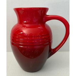 May include: A glossy, bright red ceramic pitcher featuring a wide, flared rim, a curved handle on the right side, and three horizontal embossed rings encircling the upper body. The pitcher has a rounded, bulbous shape and a smooth finish.