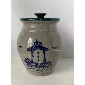 May include: A ceramic jar with a lid, featuring a lighthouse design. The jar is gray with a green rim and lid. The lighthouse is blue, with a white body and is decorated with blue birds.