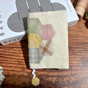 Può includere: Una copertina di libro beige con un segnalibro in tessuto. Il segnalibro è realizzato in tessuto patchwork in tonalità di verde, giallo, marrone e rosa. Il tessuto è cucito insieme in un motivo esagonale.