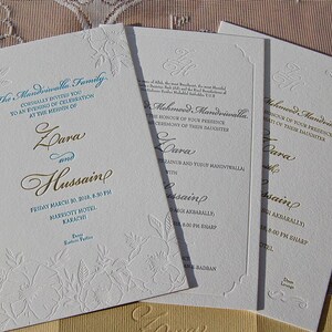 May include: Ivory wedding invitations with gold and teal text. The invitations feature floral embossed designs and include the names Zara and Hussain, along with event details. The text reads "The Mandviwalla Family".