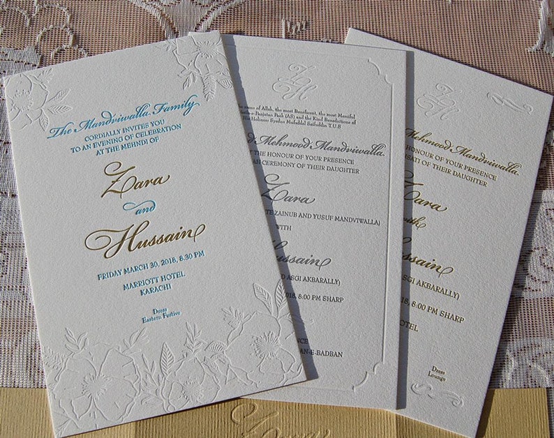 May include: Ivory wedding invitations with gold and teal text. The invitations feature floral embossed designs and include the names Zara and Hussain, along with event details. The text reads "The Mandviwalla Family".