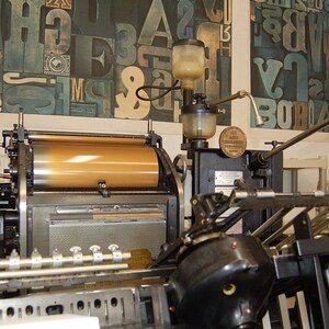 May include: An antique printing press with a large, cylindrical roller and various mechanical components. The press is black and gold, with a series of levers and gauges. The background features a wall with large, colorful letterforms.