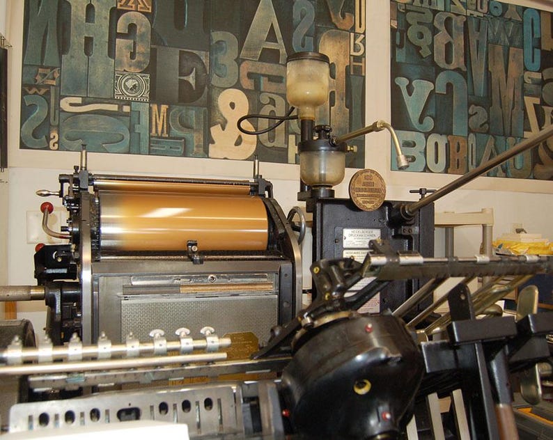 May include: An antique printing press with a large, cylindrical roller and various mechanical components. The press is black and gold, with a series of levers and gauges. The background features a wall with large, colorful letterforms.