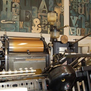 May include: A vintage printing press with a large, cylindrical roller and various mechanical components. The machine is black and gold, with a series of levers and gauges. The background features a wall with large, colorful letterforms.