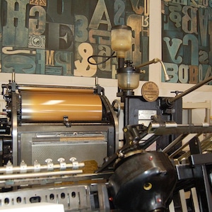May include: A vintage printing press with a large, cylindrical roller and various mechanical components. The machine is black and gold, with a series of levers and gauges. The background features a wall with large, colorful letterforms.