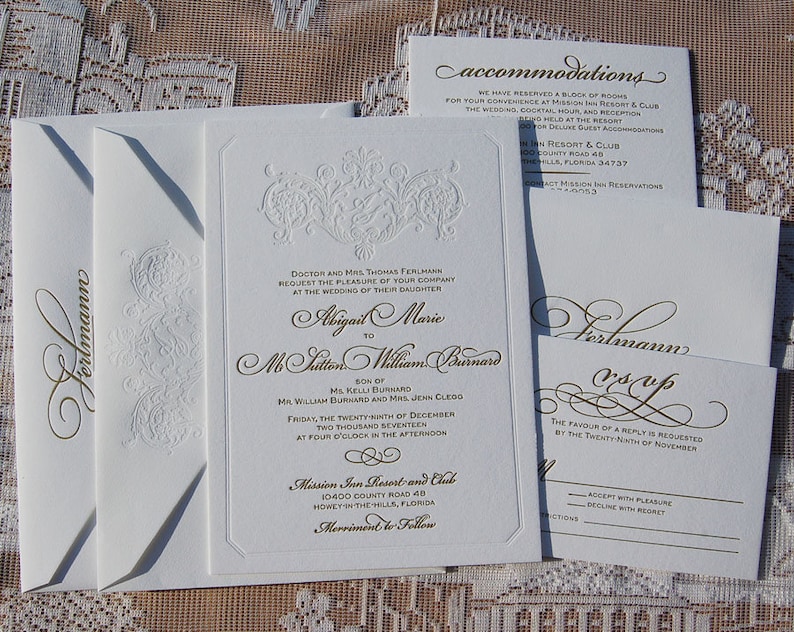 May include: Elegant white wedding invitation suite with gold script and embossed floral details. Includes invitation, RSVP card, accommodations card, and envelopes. The invitation text includes the names Abigail Marie and M. Milton William Burnard.