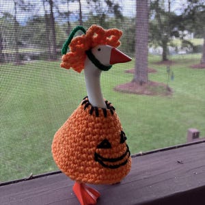 May include: A white goose figurine wearing a crocheted orange pumpkin jumper with a black stitched jack-o'-lantern face and a matching orange crocheted pumpkin hat with a green stem. The goose has an orange beak and orange webbed feet.