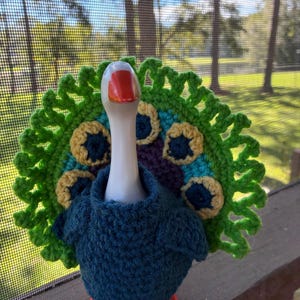 Peut inclure: Une figurine d'oie en céramique blanche portant un pull en crochet bleu foncé et une grande queue de paon en crochet circulaire et multicolore avec des accents de laine verte, jaune, bleue et violette, debout sur des pattes orange vif.
