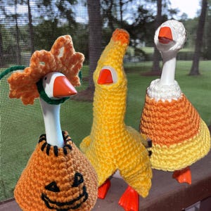 Op de afbeelding: Drie witte ganzenbeeldjes met gehaakte outfits. De eerste draagt een oranje pompoentrui en een bijpassende bloemenhoed. De tweede draagt een gele outfit met capuchon. De derde draagt een wit, oranje en geel snoepmaïs kostuum.