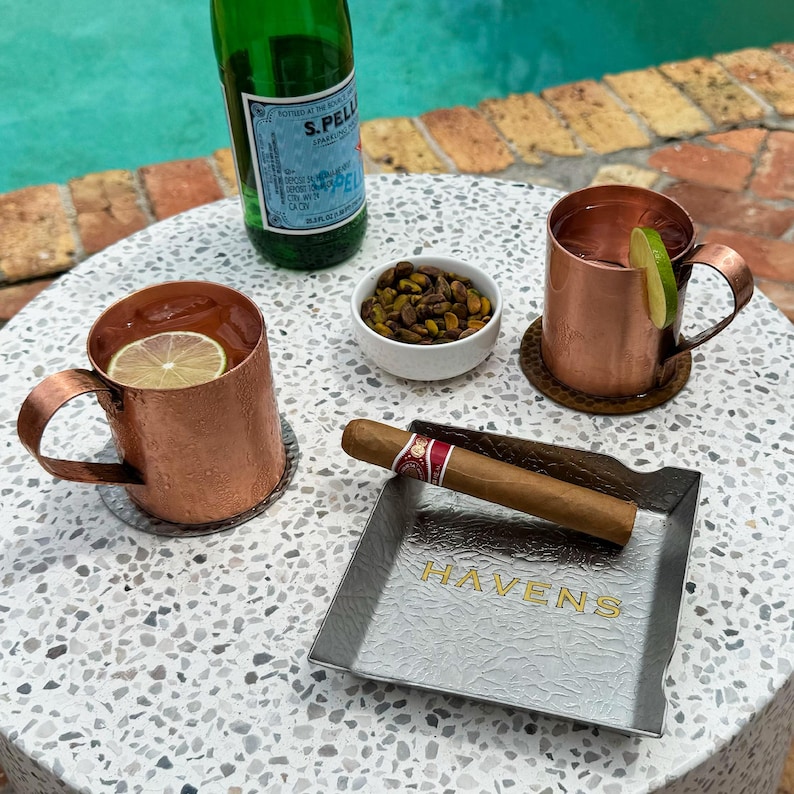 Puede incluir: Dos jarras de cobre llenas de una bebida refrescante, adornadas con rodajas de lima, se encuentran sobre una mesa con un cenicero de plata grabado con la palabra "HAVENS". Un cigarro descansa en el cenicero, y un peque&ntilde;o cuenco de pistachos se encuentra cerca.