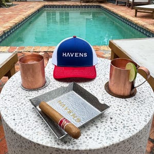 Puede incluir: Una gorra de b&eacute;isbol blanca, roja y azul con la palabra "HAVENS" bordada en la parte delantera. La gorra est&aacute; sobre una mesa junto a dos jarras de cobre, una bandeja de plata y un cigarro.