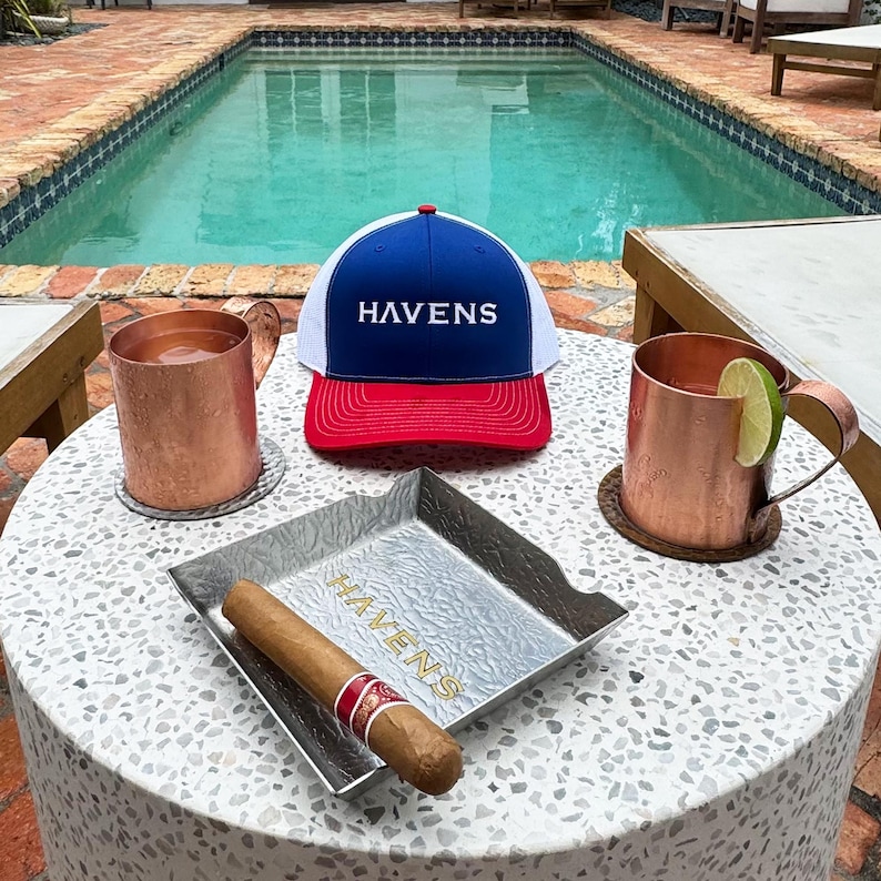 Puede incluir: Una gorra de b&eacute;isbol blanca, roja y azul con la palabra "HAVENS" bordada en la parte delantera. La gorra est&aacute; sobre una mesa junto a dos jarras de cobre, una bandeja de plata y un cigarro.