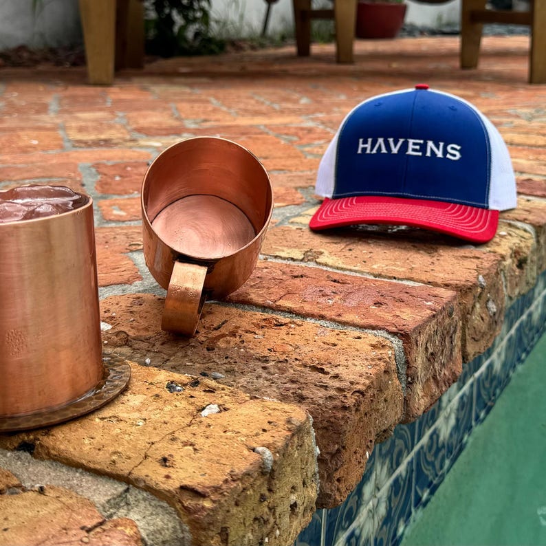 Puede incluir: Dos jarras de cobre con una gorra de b&eacute;isbol rojo, blanco y azul con la palabra "HAVENS" bordada. La gorra est&aacute; sobre una superficie de ladrillo.