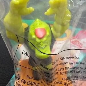 May include: A green plastic toy figure with a red nose and black hair is inside a clear plastic bag. The figure has its arms raised. The bag has text that reads "CAUTION" and "1996 Warner Bros."