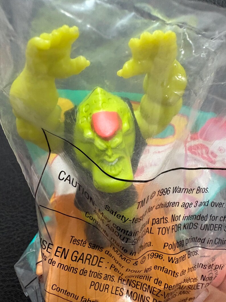 May include: A green plastic toy figure with a red nose and black hair is inside a clear plastic bag. The figure has its arms raised. The bag has text that reads "CAUTION" and "1996 Warner Bros."