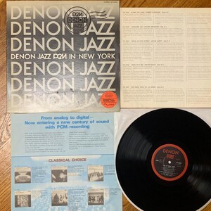 Könnte beinhalten: Eine schwarze Schallplatte mit einem roten und weißen Etikett, auf dem "Denon PCM" steht. Die Schallplatte liegt auf einer schwarzen Plattentellermatte. Das Schallplattencover ist weiß mit schwarzem Text, auf dem "Denon Jazz PCM in New York" steht.