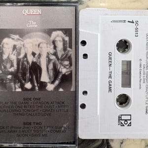 May include: A vintage cassette tape of Queen's album "The Game." The album cover features a black and white photo of the band. The cassette is white with the album title and song list printed on it. A collectible music item.
