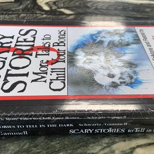 Puede incluir: Una pila de tres libros titulados "Scary Stories" de Alvin Schwartz. El libro superior tiene una cubierta roja y blanca con el t&iacute;tulo "Scary Stories: More Tales to Chill Your Bones". Los libros est&aacute;n envueltos en pl&aacute;stico transparente.
