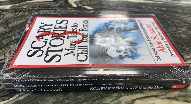 Puede incluir: Una pila de tres libros titulados "Scary Stories" de Alvin Schwartz. El libro superior tiene una cubierta roja y blanca con el t&iacute;tulo "Scary Stories: More Tales to Chill Your Bones". Los libros est&aacute;n envueltos en pl&aacute;stico transparente.