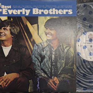 May include: Album cover for "The Very Best of The Everly Brothers" featuring a blue banner with white text. The cover shows two men smiling, with a vinyl record in a plastic sleeve to the right.