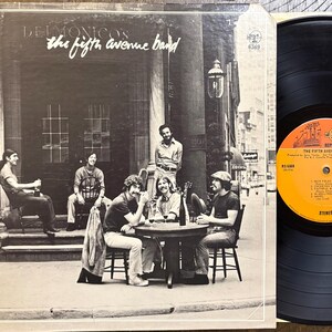 May include: Album cover for "The Fifth Avenue Band" by Delmonicos. The cover features a black and white photo of the band members sitting at a table outside a building. A vinyl record is visible on the right.