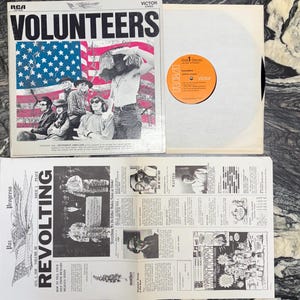 May include: Vintage vinyl record album cover for "Volunteers" by The Jefferson Airplane, featuring a group photo against a backdrop of the American flag. Includes the record and a newspaper with the headline "Revolting".