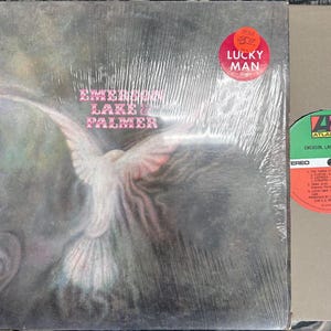May include: A sealed vinyl record album by Emerson, Lake & Palmer, titled "Lucky Man." The album cover features a dove illustration with the band's name in pink text. A red sticker with the text "Lucky Man" is on the cover. The record label is visible.
