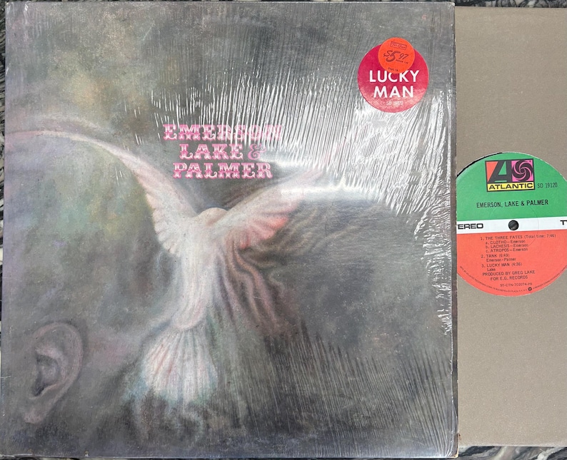 May include: A sealed vinyl record album by Emerson, Lake & Palmer, titled "Lucky Man." The album cover features a dove illustration with the band's name in pink text. A red sticker with the text "Lucky Man" is on the cover. The record label is visible.