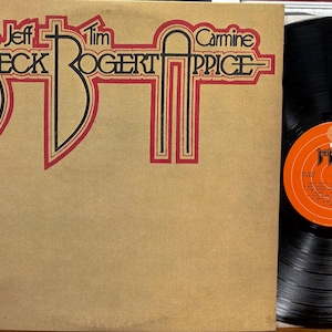 Puede incluir: Un álbum de vinilo vintage con los nombres "Jeff Beck", "Tim Bogert" y "Carmine Appice" en una fuente estilizada roja y negra sobre un fondo beige. El disco en sí es negro con una etiqueta naranja.