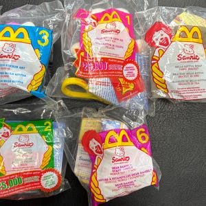 May include: A collection of sealed McDonald's Happy Meal toys featuring Hello Kitty characters. Each package is clear with colorful graphics, including the Sanrio logo and product descriptions. The toys include a sticker set, soft toy, bracelet, and mobile toy.