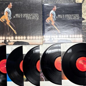 May include: Two vinyl record albums by Bruce Springsteen & The E Street Band, titled "Live/1975-85." The album covers feature a live performance image of Springsteen playing a guitar. Several black vinyl records with red labels are visible.